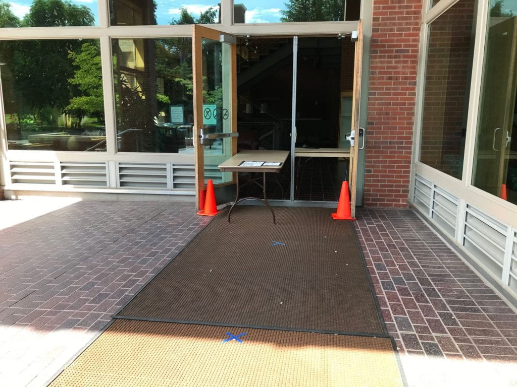 Photograph of the side door to Reid Campus Center, with a long table extending out the door, orange cones marking the doorway, and blue tape marking places to stand in line