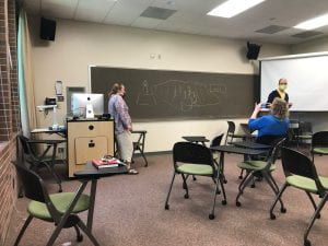 The artist stands by the chalkboard while two others gesture and talk to each other