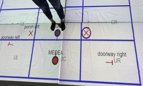 The floor-projected blocking map shows actors where to move on stage. A person stands near their mark and will next move towards the X.