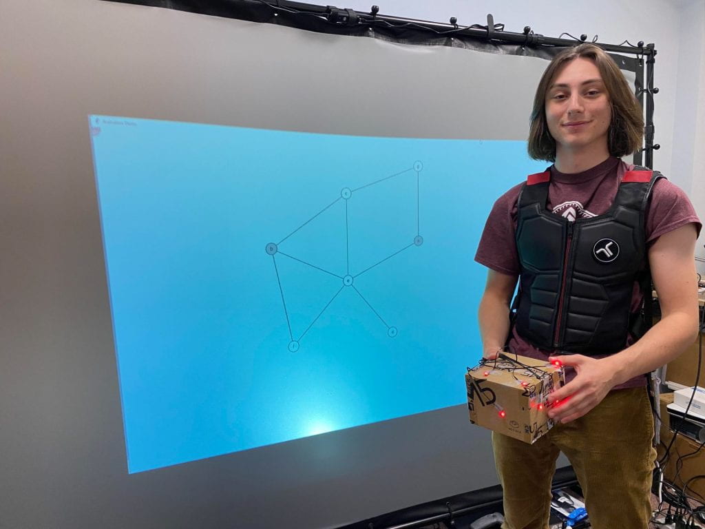 Student standing wearing a haptic vest and holding a box outfitted with red LED tracking markers. Behind the student is a rear-projection screen displaying a graph of nodes and edges to plan navigation directions.