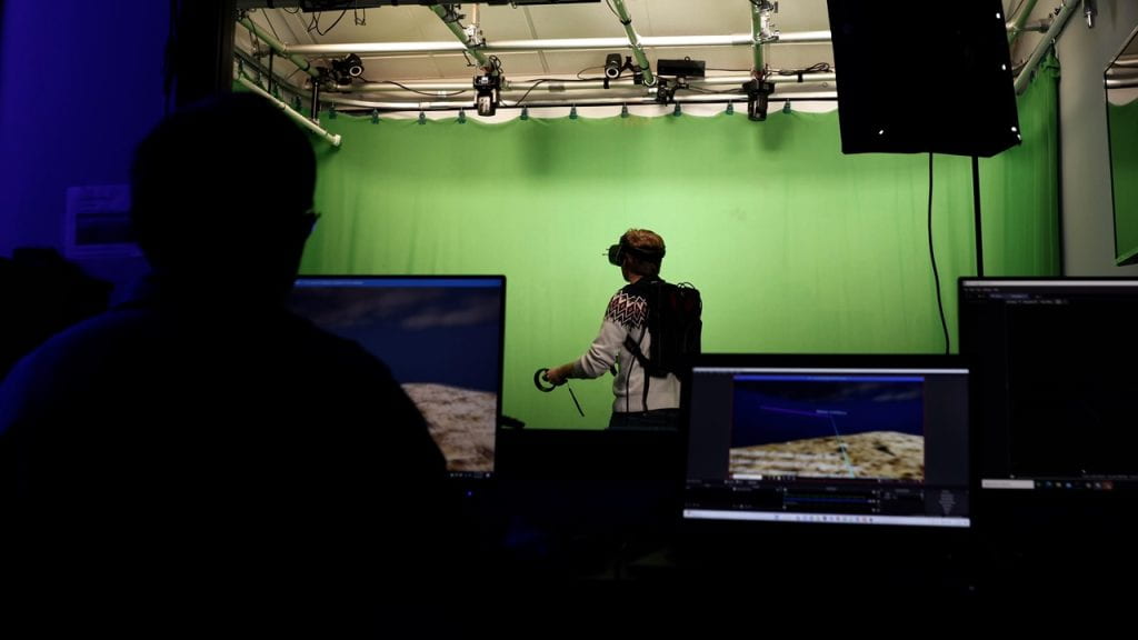 Computer Science senior Nick Hager demonstrates a VR project in the Immersive Stories Lab. Computer monitors running the geology VR program are in the foreground. Nick stands wearing a VR backpack computer, VR headset, and hand controllers. Behind Nick is the lab's green screen curtain.